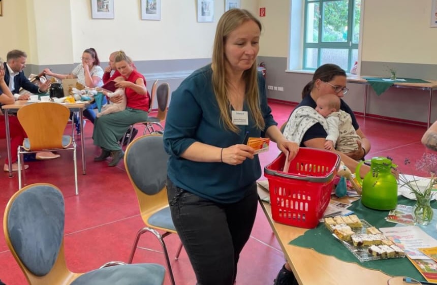 Eine Frau steht neben einen Korb, im Hintergrund sind junge Eltern mit Babies zu sehen